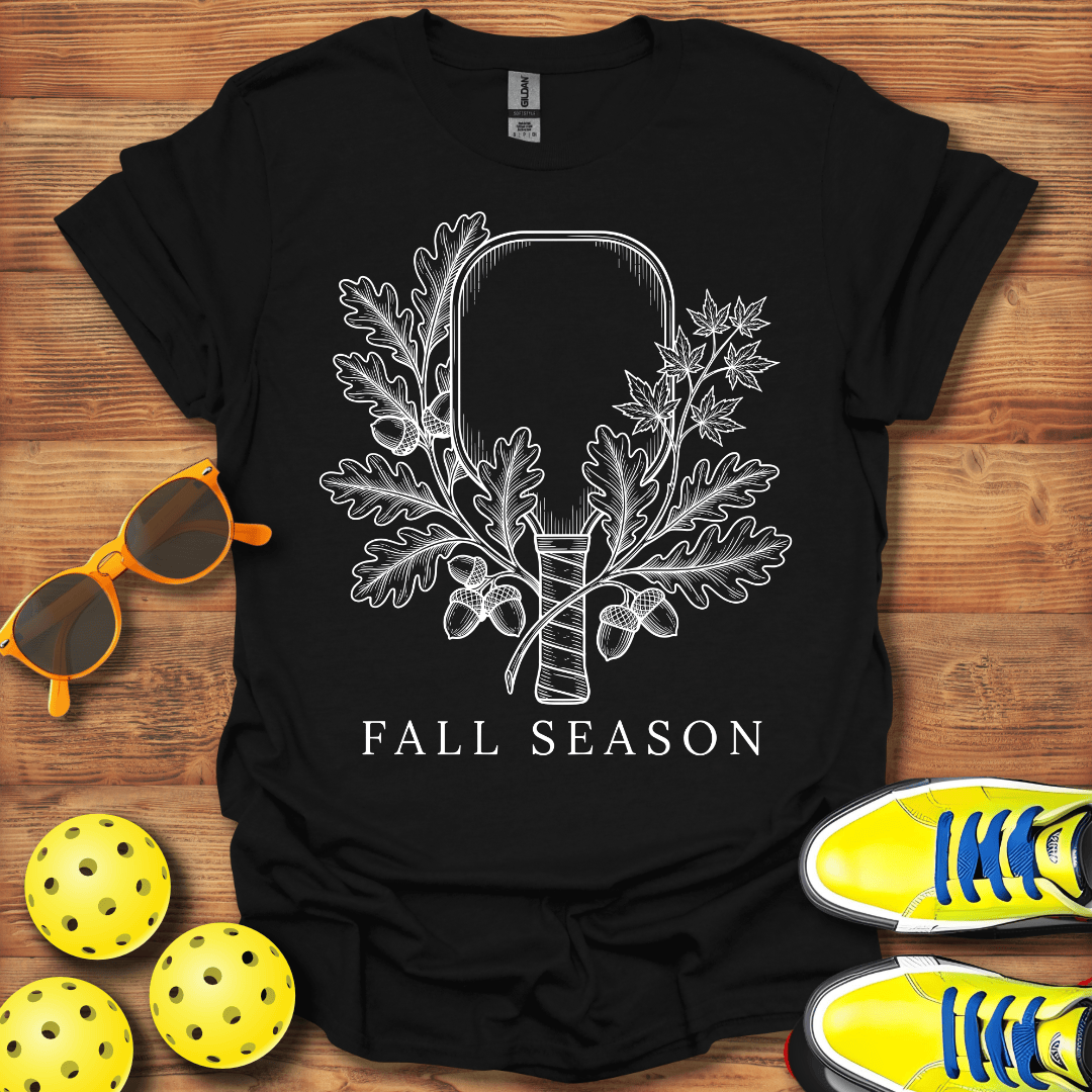 A black short-sleeve graphic T-shirt laid flat on a wooden surface, featuring a white line-art design of an axe surrounded by oak leaves and acorns with the words 'FALL SEASON' below. 