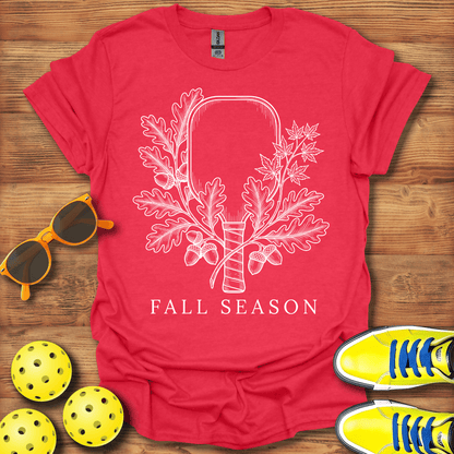 A red short-sleeve t-shirt laid flat on a wooden surface, featuring a white line-art graphic of an axe surrounded by oak leaves and acorns, with the words 'FALL SEASON' printed below. 