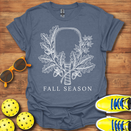 A slate blue graphic t-shirt laid flat on a wooden surface, featuring a white line art design of an axe surrounded by oak leaves and acorns with the text 'FALL SEASON'. 